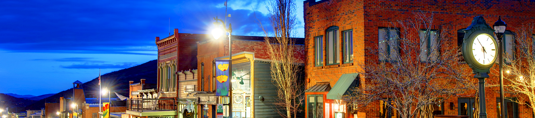 A quiet western street lined with brick storefronts, street lamps, and a clock tower, glowing under a vivid blue twilight sky.