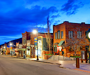 A quiet western street lined with brick storefronts, street lamps, and a clock tower, glowing under a vivid blue twilight sky.
