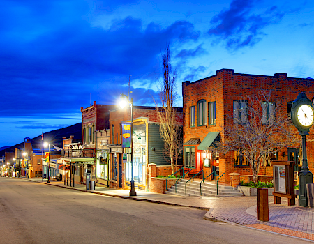 A quiet western street lined with brick storefronts, street lamps, and a clock tower, glowing under a vivid blue twilight sky.