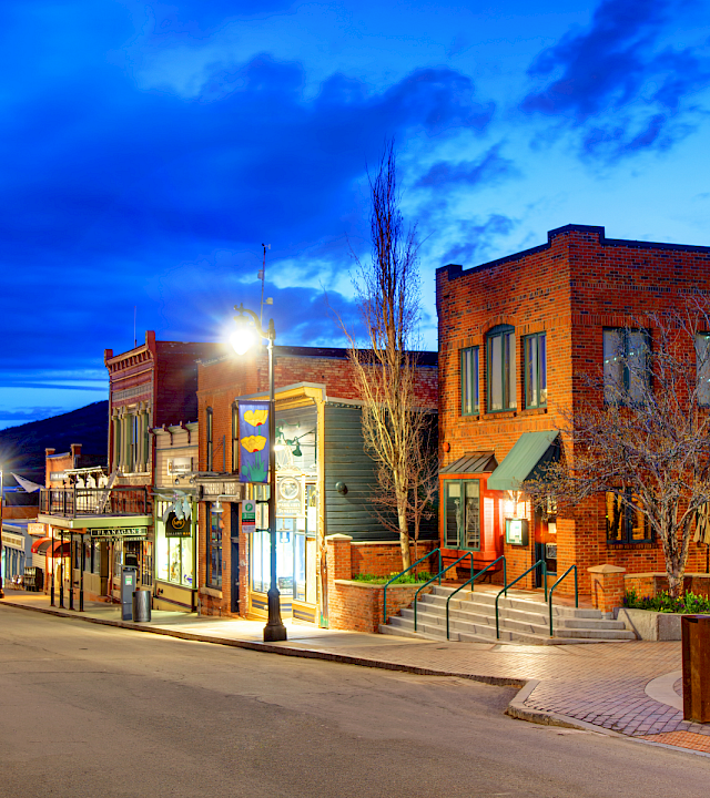 A quiet western street lined with brick storefronts, street lamps, and a clock tower, glowing under a vivid blue twilight sky.