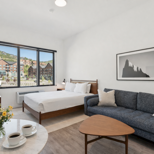 A bright hotel room with a large window, a bed, a gray sofa, a wooden coffee table, and a small round table with coffee and flowers, ending this sentence.