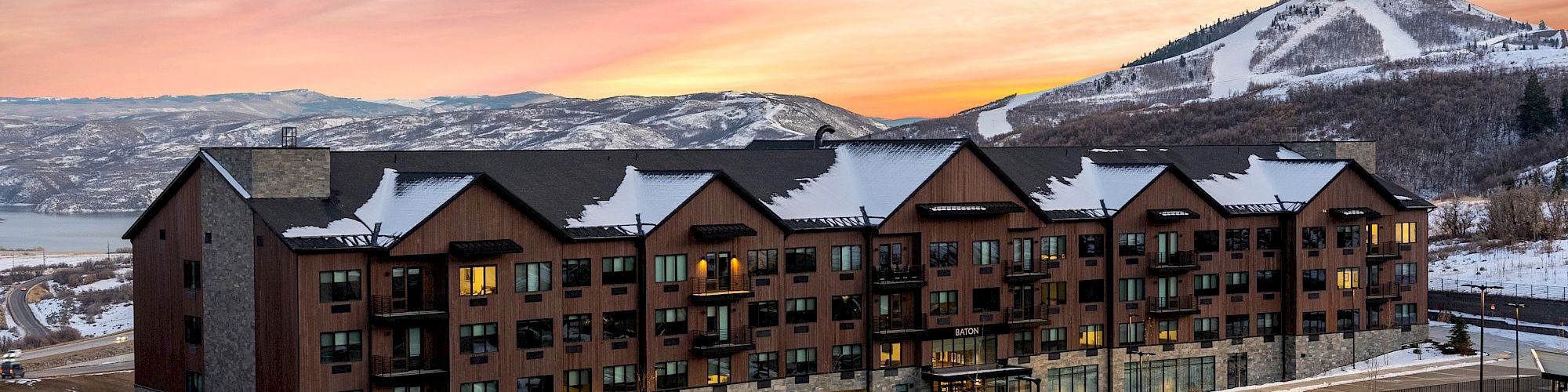 Snowy mountain sunrise over a modern hotel, with lit windows, parking lot, and hills in the distance.