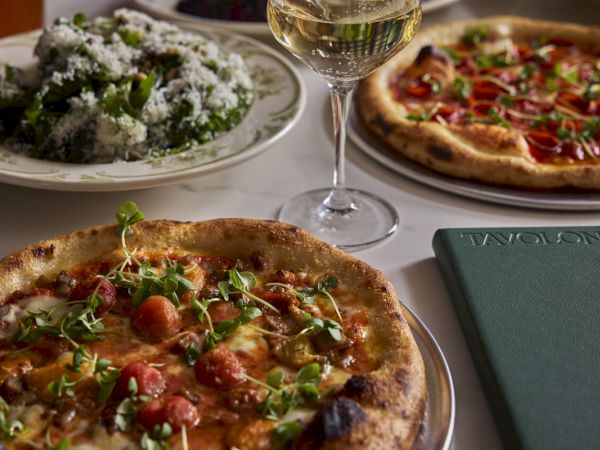 A table of pizza with microgreens, salad, and a glass of white wine beside a green book, all ready to eat.