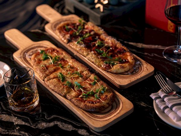 Two wood-fired pizzas on wooden boards with microgreens, served with red wine in glasses at a table.