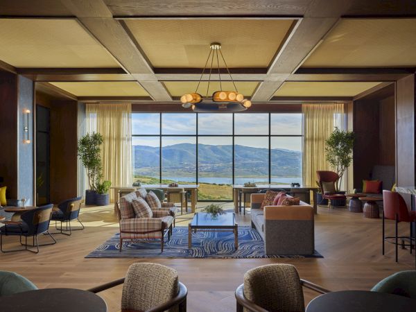 A modern lounge with sofas and a large chandelier, overlooking hills and a calm lake through huge windows.