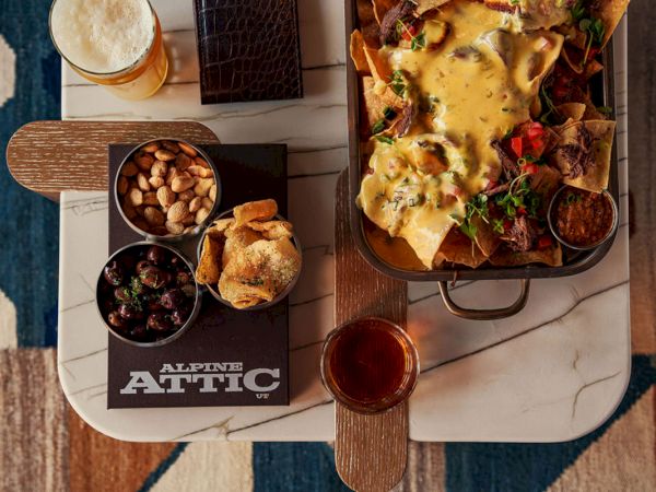 A gourmet nacho platter with melted cheese, chips, olives and nuts, plus bread and a drink on an Alpine Attic table.