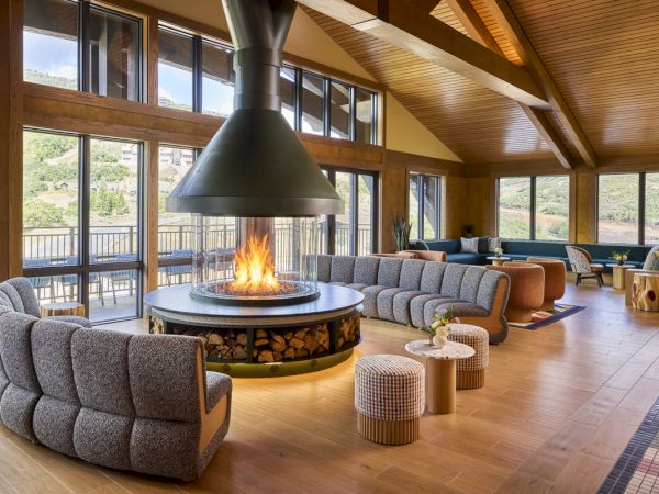 A cozy modern lodge living room with large windows and a roaring circular fireplace, plush seating, and warm wooden beams overhead.
