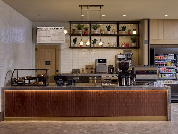 A modern coffee shop counter with espresso machines, pastries in a display, and a stocked drink fridge.