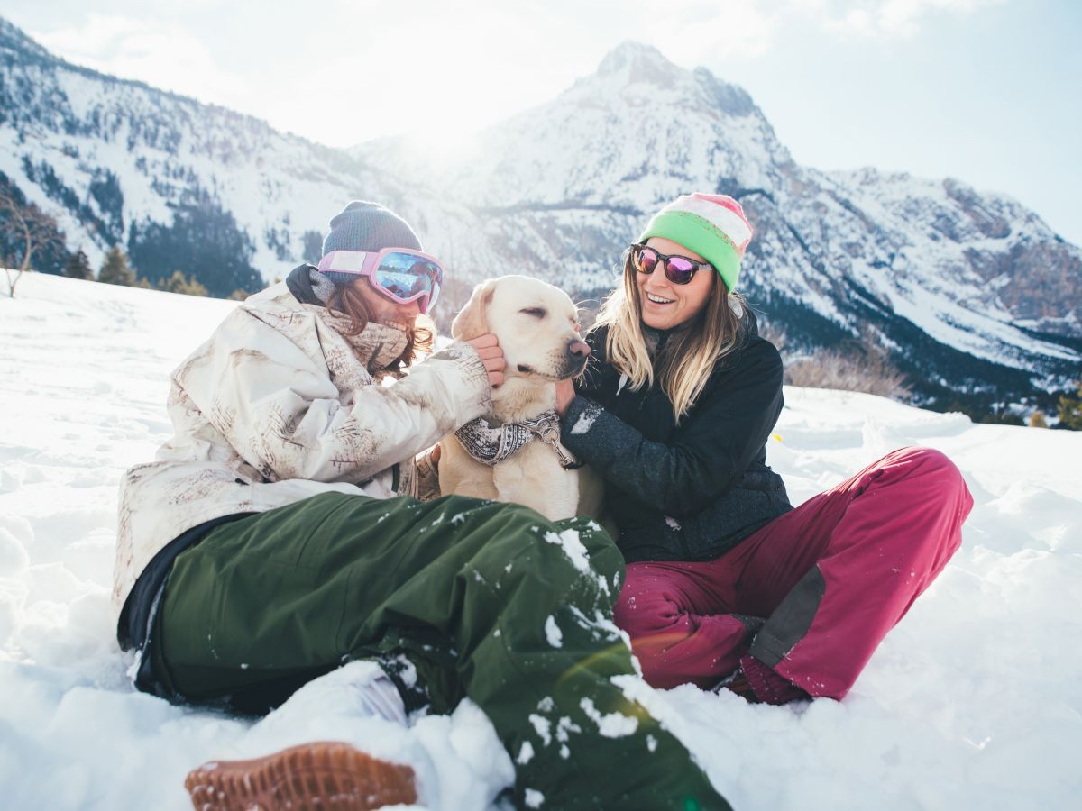 In Utah mountains, a winter couple hugs a dog while wearing goggles and beanies, with mountains in the background.